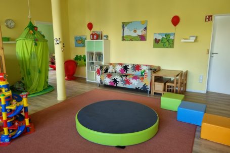 Heller, freundlicher Spielraum mit gelben Wänden, einem grünen Kuschelzelt, einer bunten Sitzecke, einem grünen Spielpodest und Bausteinen.